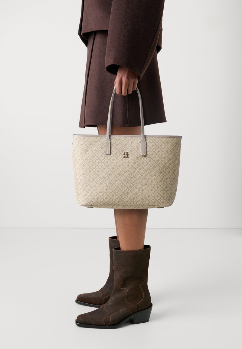 Borsa a tracolla beige con motivo e manici in pelle grigia, con un logo in evidenza. Il modello indossa un abito marrone e stivali marroni abbinati.