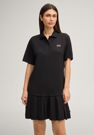 Frau, die ein schwarzes Polo-Kleid mit kurzen Ärmeln und plissiertem Rock trägt, mit dem "JOOP! JEANS" Logo auf der Brust, vor einem einfarbigen Hintergrund stehende.
