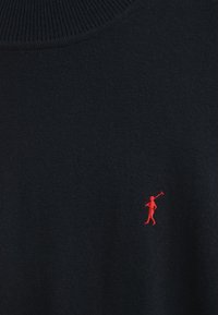 Marinblå ulltröja med ribbad krage, prydd med en liten röd broderad figur på vänster bröst. Slät textur genom hela tröjan.