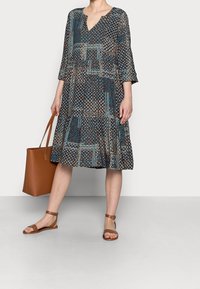 Vestido impreso hasta la rodilla con manga tres cuartos en tonos azul marino, marrón y beige. Presenta un escote en V y dobladillo acampanado. Bolso tote marrón y sandalias.