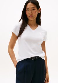 Ženska z dolgimi temnimi lasmi, oblečena v belo majico z V-izrezom in temno modre hlače s črnim pasom, stoji z eno roko v žepu.