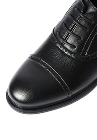 Scarpa da uomo in pelle nera con punta a cappuccio, dettagli brogue e allacciatura chiusa su sfondo bianco.