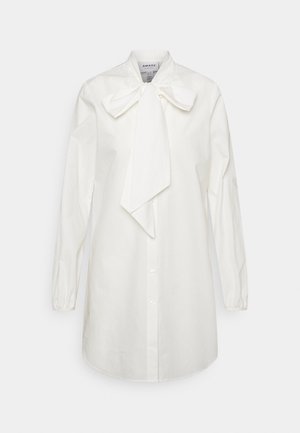 Longue blouse blanche boutonnée à manches longues avec un grand nœud décoratif noué au col.