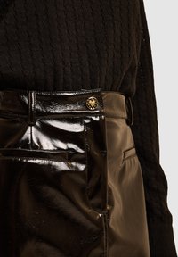 Pantaloni in pelle verniciata nera con una finitura lucida, caratterizzati da un dettaglio di bottoni dorati e una tasca frontale, abbinati a un top nero testurizzato.