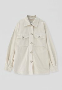 Chemise beige à manches longues avec boutons, deux poches à rabat à l'avant et un col classique, étalée à plat sur un fond clair.