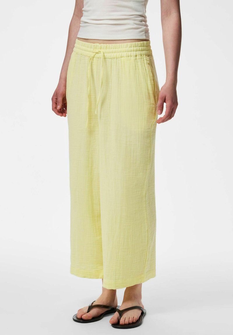 Personne portant un pantalon large ample jaune clair avec une taille à cordon et des tongs noires, un crop top blanc partiellement visible.