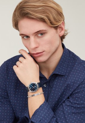 Giovane uomo con capelli biondi che indossa una camicia blu con motivi, mostrando un orologio da polso argento e blu e un braccialetto d'argento sul polso sinistro.