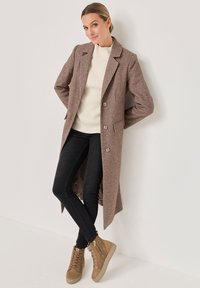 Brauner Wollmantel mit strukturierter Oberfläche, zwei Knöpfen und Seitentaschen, getragen über einem weißen gerippten Pullover und schwarzen Jeans, kombiniert mit braunen Stiefeln.