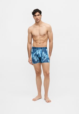 Bărbat fără cămașă, desculț, purtând chiloți boxer albaștri cu model și talie „BJÖRN BORG” pe fundal alb simplu.