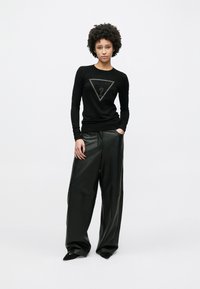 Μαύρο πουλόβερ με μακριά μανίκια και τριγωνικό λογότυπο "GUESS?" στο κέντρο, συνδυασμένο με υπερμεγέθη μαύρο δερμάτινο παντελόνι. Λείο υφή.