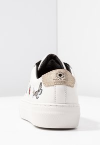 Bílé tenisky s vyšitými motýli a hmyzem, s texturovaným zlatým akcentem na patě a černým vnitřním podšívkem. Plochá podešev.