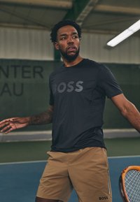 Fekete, nedvességel elvezeto sportpóló "BOSS" logóval. Bézs rövidnadrághoz párosítva, oldalsó zsebbel és logóval a szegélyen. Teniszpálya háttér.