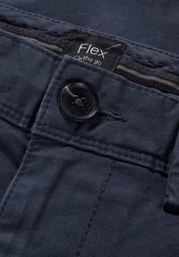 Gros plan sur la ceinture d’un pantalon bleu foncé montrant un bouton noir, une boutonnière, un passant de ceinture et une étiquette indiquant "Flex on the go."