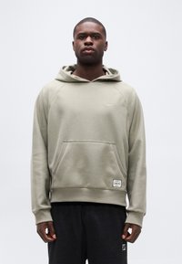 Μαύρος άνδρας που στέκεται, φορώντας ανοιχτό γκρι φούτερ Nike με τσέπη μπροστά και σκούρο παντελόνι, σε απλό λευκό φόντο.