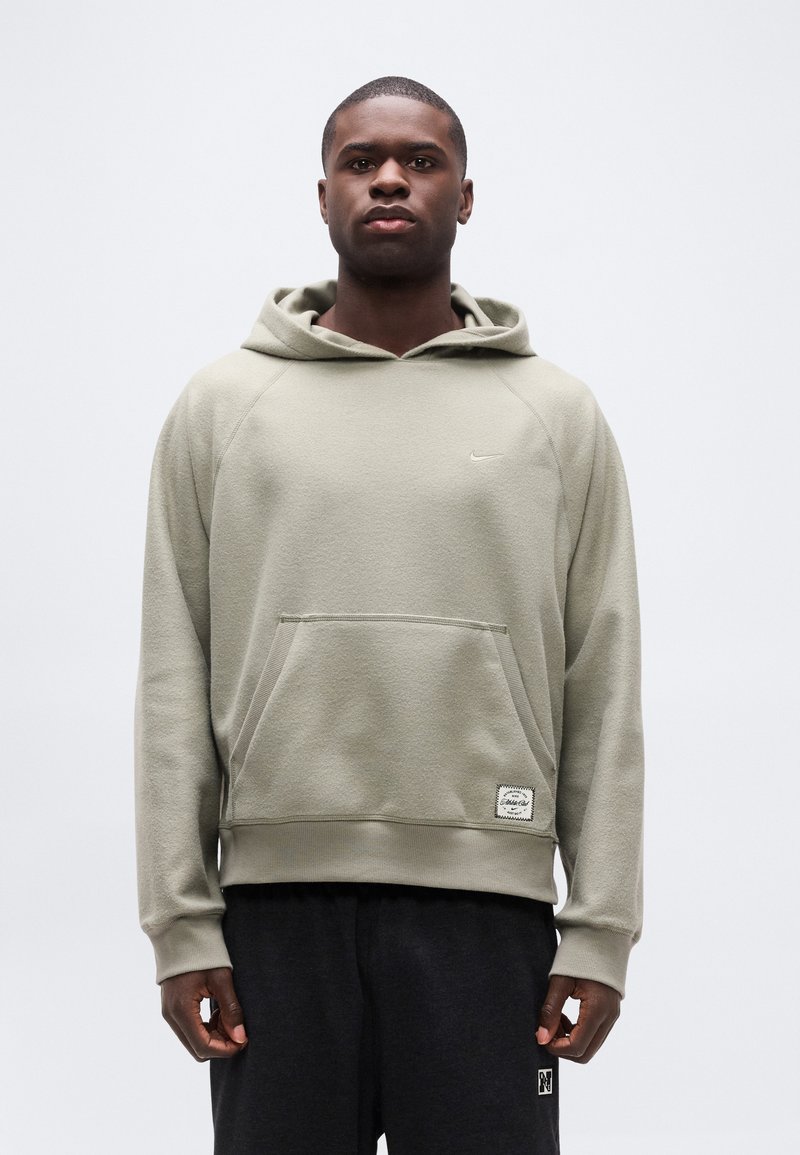 Μαύρος άνδρας που στέκεται, φορώντας ανοιχτό γκρι φούτερ Nike με τσέπη μπροστά και σκούρο παντελόνι, σε απλό λευκό φόντο.