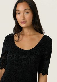 Musta pitsitoppi, jossa on lyhyet hihat, pyöreä kaula-aukko ja kukkakuviot. Rypytetty ja puoliksi läpinäkyvä kangas, jossa on aaltoilevat reunat.