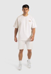 Camiseta de algodón de color beige claro con mangas cortas y escote redondo, combinada con pantalones cortos a juego. Presenta un pequeño logo en el pecho.