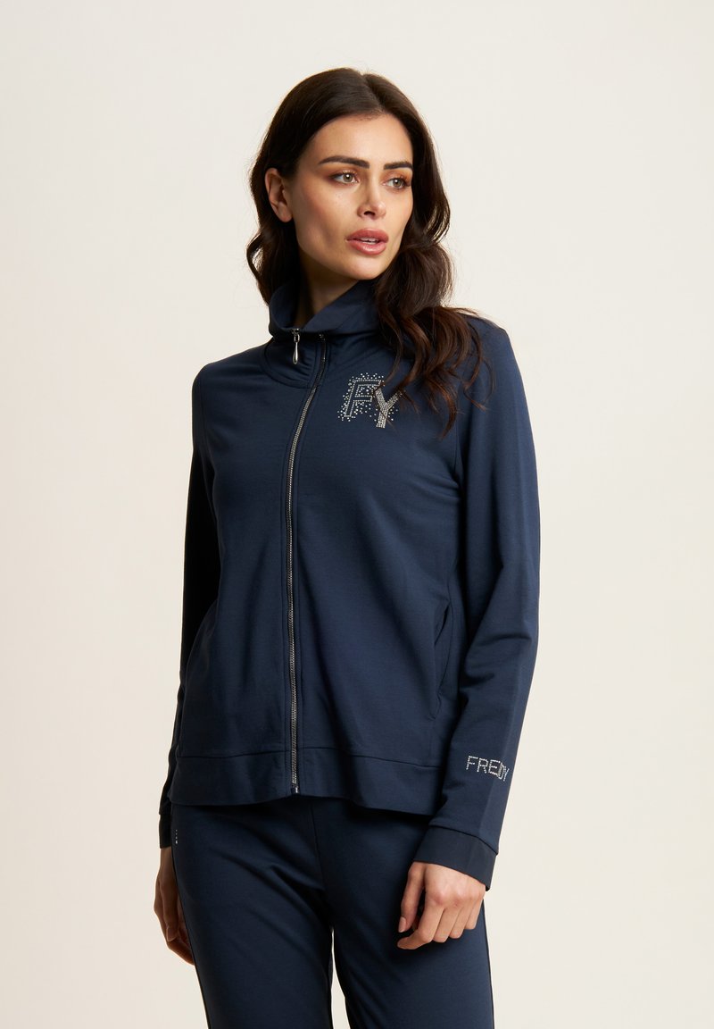 Giacca zip in blu navy realizzata in morbido tessuto con colletto alzato. Presenta un logo in strass sul petto e un dettaglio del logo sui polsini. Progettata per il massimo comfort.