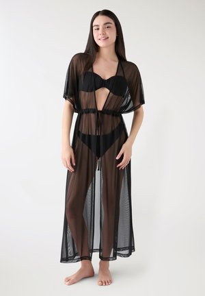 Donna che indossa un copricostume in rete nera trasparente annodato in vita sopra un bikini nero, in piedi a piedi nudi su sfondo bianco.