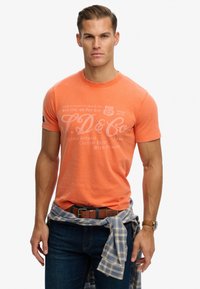 Camiseta de algodón naranja con logo y texto impresos en blanco. Diseño ajustado, mangas cortas, combinada con jeans oscuros y una camisa de cuadros azul atada a la cintura.