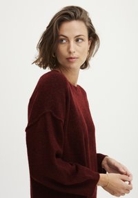 Maglione in lana bordeaux con una vestibilità rilassata, caratterizzato da spalle abbassate e polsini a costine, mostrato su un modello che si trova leggermente di lato.