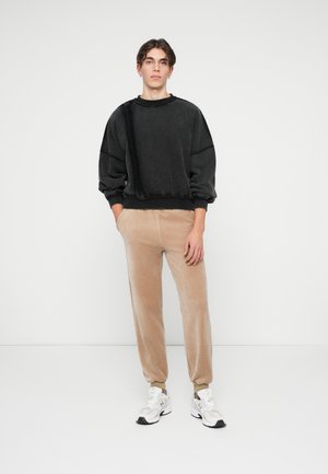 Jonge man staat met zijn handen in zijn zakken, gekleed in een zwart oversized sweatshirt, beige joggerbroeken en witte sneakers tegen een witte achtergrond.