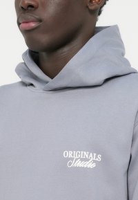 Ανοιχτό γκρι φούτερ με κουκούλα από απαλό ύφασμα, με λευκό λογότυπο «ORIGINALS Studio» στο στήθος. Απλός σχεδιασμός με άνετη εφαρμογή.