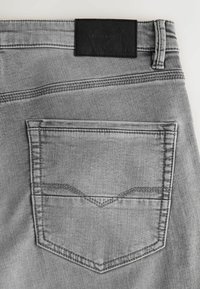 Blugi gri din denim, cu un buzunar spate ce are detalii cusute, o plasture de piele neagră texturată și variații subtile în finisajul țesăturii.