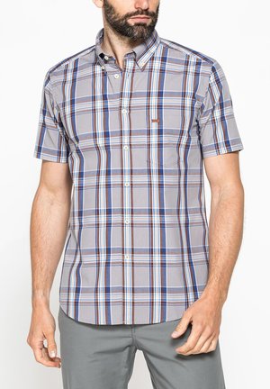 Uomo che indossa una camicia a maniche corte a quadri blu, marrone e bianco e pantaloni grigi, in piedi di fronte a uno sfondo bianco.