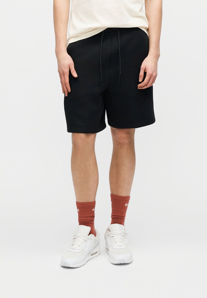 Personne portant un short noir à cordon, des chaussettes rouges Nike, des baskets blanches et une chemise beige, debout devant un fond blanc.