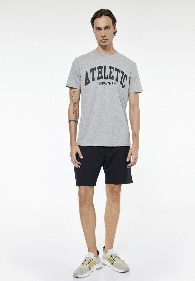 Young man with tattoos wearing a grey "ATHLETIC DEPARTMAN" t-shirt, black shorts, and beige sports shoes standing against a white background.