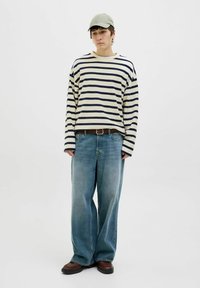 Camisa de rayas en crema y azul marino de manga larga, jeans azules holgados, cinturón marrón, zapatos marrones, gorra verde claro, de pie sobre un fondo neutro.