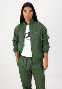 Grüne gesteppte Jacke mit hohem Kragen, Reißverschluss und elastischen Bündchen, kombiniert mit passenden Hosen, die eine subtile strukturierte Textur aufweisen.