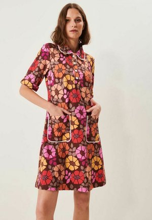 Kvinde iført en brun kjole med store lyserøde, røde, orange og gule blomster, forlommer og hvide kantninger på krave og lommer.