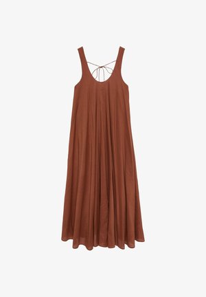 Femme portant une robe maxi sans manches couleur rouille avec dos ouvert et détail à nouer, marchant avec des chaussures noires ouvertes à l’arrière sur fond gris.