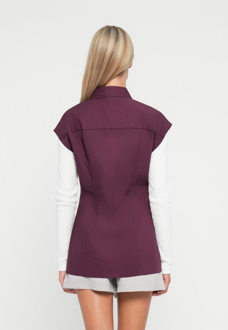 Donkerpaarse op maat gemaakte blazer met korte mouwen, aansluitend ontwerp en een kraag. Draagt over een witte blouse met lange mouwen en grijze shorts.