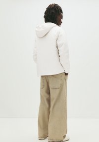 Person mit Dreadlocks, der eine weiße Kapuzenjacke, beige weite Hosen und weiße Sneakers trägt, steht mit dem Rücken zu einer schlichten weißen Fläche.