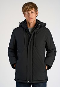 Giacca imbottita nera con cappuccio removibile, due tasche frontali, chiusura con zip e polsini elastici, indossata sopra un maglione grigio.