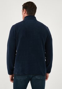 Giacca in pile blu navy, texture morbida, colletto alto, maniche lunghe e orlo nero a contrasto. Indossata sopra jeans in denim scuro.