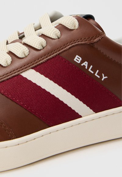 Barna bőr sportcipő bordó vászon csíkkal, krém színű fűzőkkel és fehér gumi talpal. A "BALLY" márkaneve fehér színben feltűnő.
