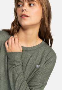 Green long-sleeve shirt made of lightweight fabric; features a round neck, cuffed sleeves, and a small branded accent on the chest.