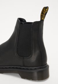 Zwart leren Chelsea boot met een gestructureerd elastisch paneel, stevige zwarte zool en een gele hieltrek met branding.