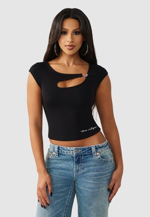Vrouw met een zwart cropped top met een sleutelgat uitsnede en gesp, gecombineerd met lichtblauwe high-waisted jeans, staand tegen een effen achtergrond.