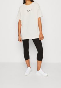 Krämfärgad T-shirt med kort ärm och svart Nike-logotyp, ihopparad med svarta caprilösa leggings och vita sneakers mot en neutral bakgrund.