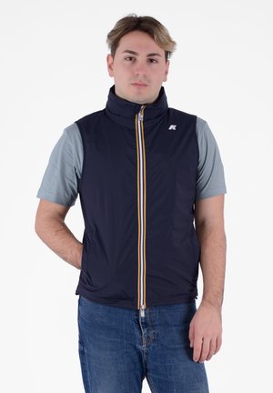 Gilet blu navy con collo alto, zip frontale gialla e bianca, tessuto leggero. Indossato sopra una camicia azzurra a maniche corte, abbinato a jeans blu.
