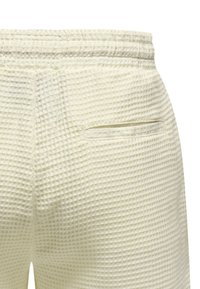 Cremefarbene, strukturierte Hose mit geometrischem Muster, ausgestattet mit einer Seitentasche und einem elastischen Bund. Der Stoff wirkt leicht und atmungsaktiv.