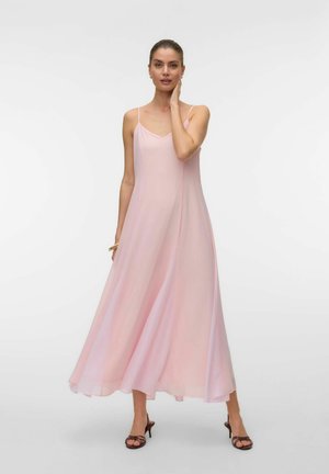 Kobieta w lejącej, jasno-różowej sukience maxi na cienkich ramiączkach i czarnych sandałkach na paski, pozująca z jedną ręką dotykającą szyi.