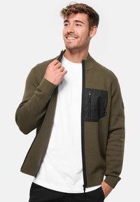 Cárdigan de cremallera en verde oliva con un cuerpo de punto texturizado, bolsillo en el pecho negro y puños acanalados, usado sobre una camiseta blanca.