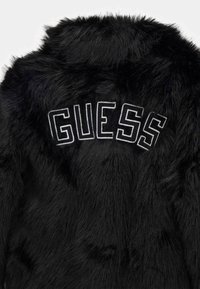 Melnā mākslīgā kažokā ar mīkstu tekstūru. Uz muguras ir drosmīgs balts "GUESS" izšuvums blokveida fontā.