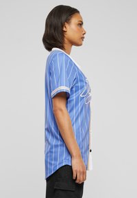 Camicia blu in stile baseball con righe verticali bianche, bottoni bianchi a contrasto e orlo arrotondato. Maniche corte con bordo bianco.